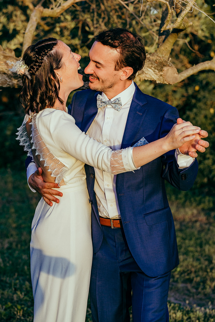un homme en costume danse avec une femme en robe de mariée blanche, la photo est prise au coucher du soleil devant un olivier par un photographe mariage montpellier